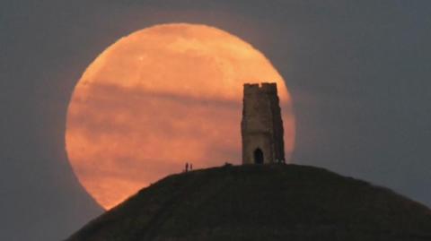 A reddish looking full moon behind a structure on a hill