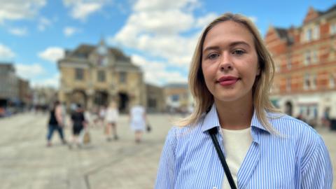 Daisy Blakemore-Creedon in front of Peterborough's Guildhall, with the background blurred out. She has shoulder-length blonde hair and is wearing pink lipstick. She has a blue and white striped overshirt on, which is above a white top. A black bag strap is visible across her chest.