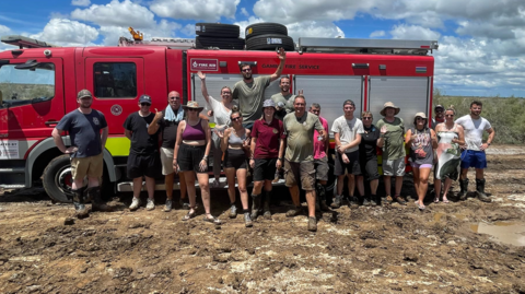 A number of people in hats and shorts stood in front of a fire engine on a muddy road