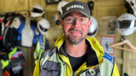 Chris Ganley stands smiling at the camera. He is wearing a black cap with the word "Yamaha" on it in white lettering. He has a dark beard and is wearing a yellow high-viz jacket