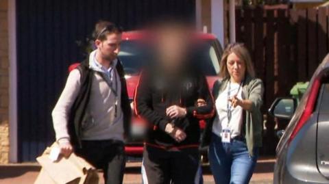 Two plain clothed detectives lead away a man into an unmarked police car in a suburban housing estate. The officers - one male, one female are in jeans and trainers. The suspect is in trainers and tracksuit bottoms and has been blurred to hide his identity.