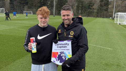 A boy with ginger hair, wearing a Nike tick black jumper and grey joggers, stood next to a man with greying black hair wearing a Wolverhampton Wanders black jacket, holding up a community captain award.