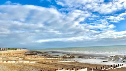 shingle beach with groynes, waves lapping on shore under a cloudy, blue sky