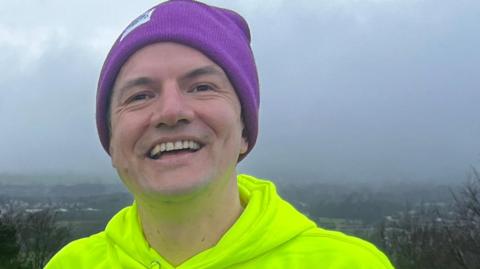 A man wearing a neon green hoodie and purple beanie hat smiles at the camera