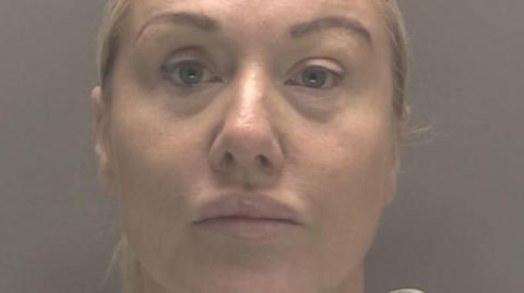 Lindy Leah in a police mugshot. She is staring, unsmiling, at the camera. She has blonde hair which is tied back and blue eyes. The background is grey.