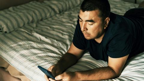 A man is lying comfortably on a striped bed, focused on browsing or reading on his smartphone. The casual setting expresses relaxation, focus, and a solitary moment.