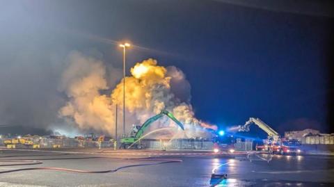 A large amount of smoke can seen coming from an area, with two cranes spraying water on the affected area. There is a fire engine to the right, and it is night-time. 