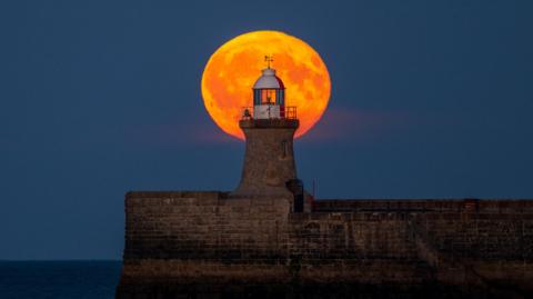 The moon is illuminated orange behind the red and white lighthouse at the end of the pier. 