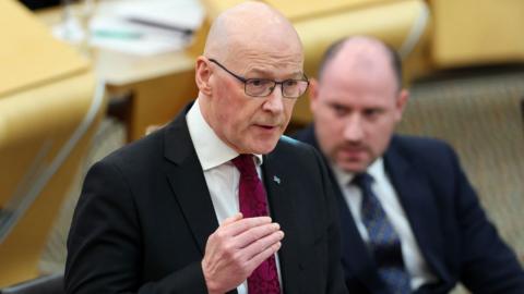 John Swinney speaking in the Scottish Parliament. He is visible from the chest up, with his right hand raised in front of his body. He is bald and is wearing a black suit, a white shirt a purple tie and glasses. 