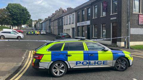 A police car parked across the road with a street with a row of houses behind it. There is police tape across the road and another police car in the background