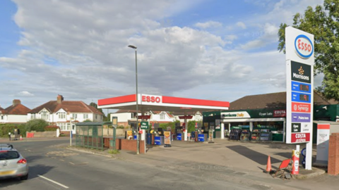 An Esso fuel station forecourt with a Morrisons Daily store. The petrol station is situated on the corner of two roads. There are no cars in the forecourt and houses can be seen on the other side of the road. It is a partly cloudy day with patches of blue sky.