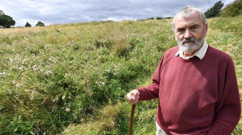 A grey-haired, bearded man with a maroon jumper and a serious expression and holding a walking stick or staff, with a field of overgrown thistles and grass in the background.