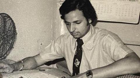 Rajinder Verma in a black and white picture of him as a young man, sitting at a desk with a newspaper. He is wearing a short-sleeved shirt with a tie, a bracelet and watch, and has fan to one side of the table. There is a calendar behind him on the wall.