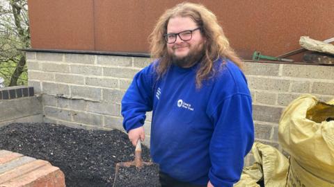 A man in a blue sweatshirt holds a hand shovel full of grey and black ash.