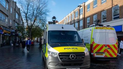Police vans mounted with facial recognition cameras are parked in a shopping street on a bright winter's day.