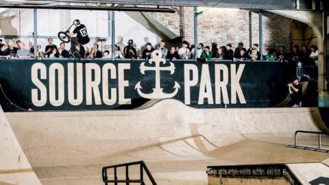 An indoor skatepark at The Source Park in Hastings with ramps, rails and BMX riders. Large "SOURCE PARK" branding is displayed above the course, with spectators gathered around the edge of the venue.