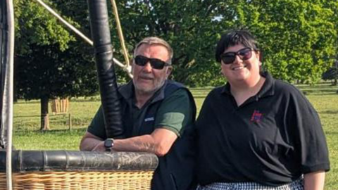 Lorna Hankey wearing a black polo top and checked trousers is standing next to a balloonist who is wearing a dark green polo top and blue gilet. The pair of them are standing next to a balloon basket.