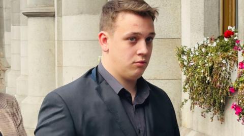 A young man with short, light-brown hair, wearing a suit jacket. He is entering a court