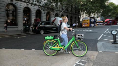 A woman wheels a green e-bike across a road in London