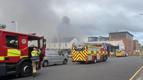 Three fire engines and several firefighters in the street next to the fire