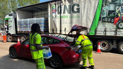 Two people in hi-vis jackets getting water bottles out of the back of a red car. There is a large lorry next to the car.