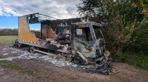 The burned shell of a lorry on a muddy surface with a field in the background