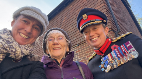 A picture of Clare Phillips in her Army uniform, with a host of medals, stood next to her mother Rhoda, who is wearing a purple coat and woolly hat, is wearing glasses and has white hair, and her sister Ali on the left. She is wearing a woolly cat, a scarf and a black coat. They are all smiling.