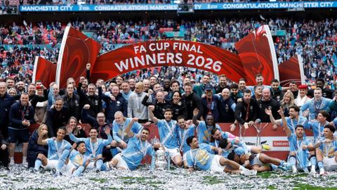 Manchester City players, manager and staff celebrate winning the FA Cup. They have the trophy on the pitch with a red sign in the backdrop which says FA Cup Final Winners 2026. 