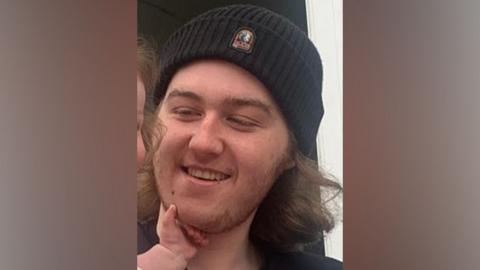 Ellis Cox has mid length brown hair and is wearing a black wooly hat. He is holding a baby and looking and smiling at her.