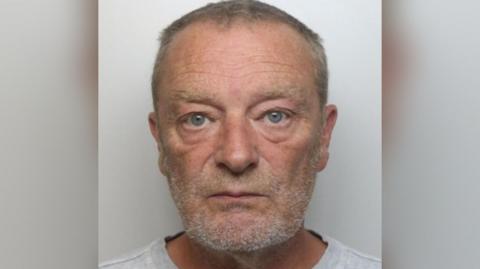 Paul Renwick looking at the camera in a police mugshot. He is an older man with short, graying hair and a beard or stubble. 