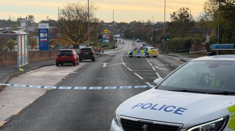 A long road stretches away. in the foreground is a police car and a cordon across the road. A bus stop on the left and two cars. Tall signs for Tesco and Lidl further down the road.