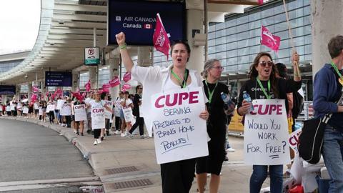 A photo showing Air Canada flight attendants picketing outside Toronto Pearson International Airport. The photo shows a long line of picketers holding signs, with a focus on two at the front with signs that read "serving pretzels to bring home peanuts" and "would you work for free?". Others are seen waving pink CUPE flags - the union representing Air Canada cabin crew.