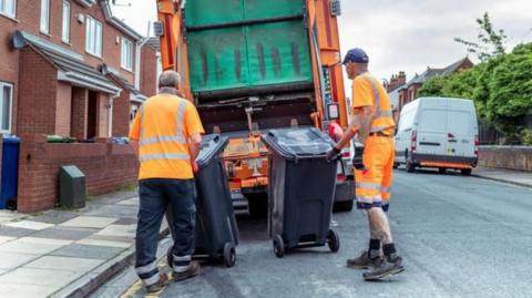 Two workers holding bins in front of a rubbish lorry in front of houses in a street. The bins are on balancing on wheels.