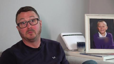 Dale Gardiner sits on a grey sofa during an interview, wearing glasses and a dark top, with a framed school photograph and a small white device on a table behind them.