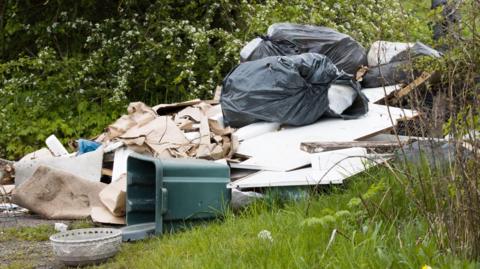 A pile of rubbish dumped in a bit of countryside. There are large bits of white MDF wood, with large blacksacks filled with rubbish, cuttings of woods, a rolled up beige carpet and a tipped over green bin. They are surrounded by grass and hedging with white flowers in the hedges. 