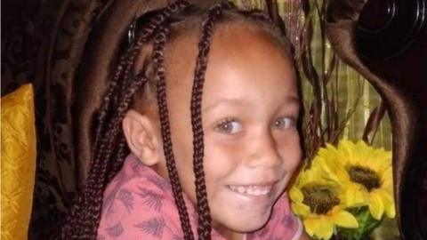 Joshlin Smith with braids smiles at the camera. She is wearing a pink top and sunflowers can be seen beside her.