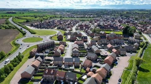 Drone footage of a new build housing estate. There is a large roundabout to the left and about 100 houses in the development.