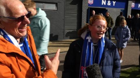 An older man in an orange jacket and Ipswich Town's blue underneath, stands facing a reporter who is off camera. He is pointing to a young woman with red hair beside him, who is in a navy coat.