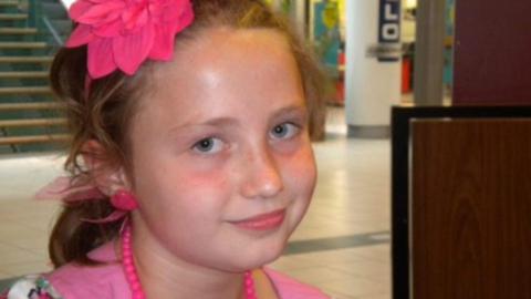 Lucie Southall. A young girl wearing a pink top and plastic pink necklace. She also has toy plastic earrings in and a pink flower. She is smiling and looking at the camera. 