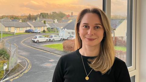 A head-and-shoulder image of Rosie Richardson. She has shoulder-length blonde hair and blue eyes. She is wearing a black jumper and a golden chain with a round pendant. She is standing in front of an office window at sunset. It looks out to a housing estate with Cockermouth Castle and Jennings Brewery visible in the skyline.