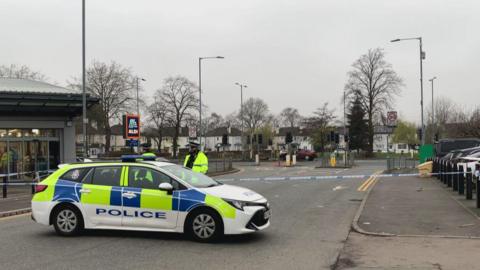 A police car is parked near the cordon which is next to the Aldi supermarket in Burnage