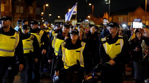 Numerous police officers in caps and hi-vis tabards stand in front of protesters carrying placards and Israeli flags on a city street at night. Some of the placards read "Keep antisemitism out of football".
