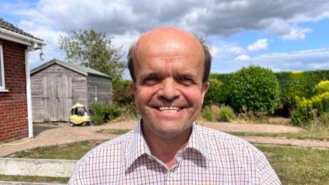 A bald smiling man in a tattersall checked shirt is standing in a garden, there's a wooden shed behind him and a child's toy car parked in front of it.