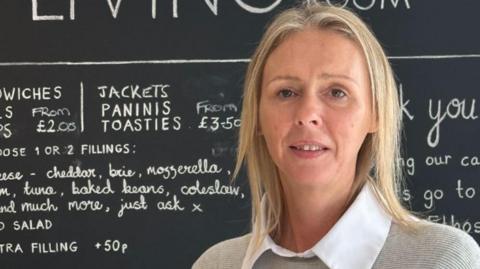 A woman with blond hair in a grey top and white blouse, stood in front of a cafe blackboard