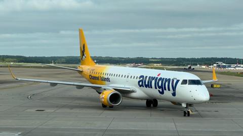A shot of an Aurigny aeroplane at an airport. It is mainly white with the company branding on it with the back of it yellow. It has Aurigny and the Channel Islands labelled on it. 