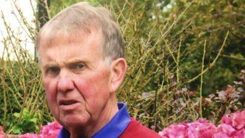 Malcolm Flynn. He is an older man with light hair. He is wearing a blue-collared shirt under a red jumper. He is standing in a garden surrounded by pink flowers and greenery.