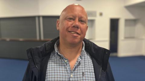 Ross Le Brun. He is wearing a checked shirt and black jacket. He is bald and smiling at the camera. 