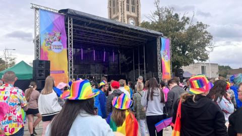 People wait for the action to start in front of a large stage. They are wearing bright clothes and rainbow hats.