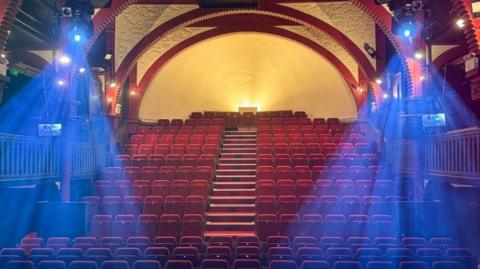 A view from the stage inside the Cheltenham Playhouse theatre, showing rows of red theatre seats with a staircase up the middle. The building has arched red beams and blue lights shining on the stage from either side of the ceiling.