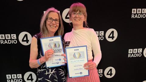 Two women are standing side by side holding up certificates and a glass award. They are both smiling. They are in front of a black back drop with the BBC Radio 4 logo printed on it.